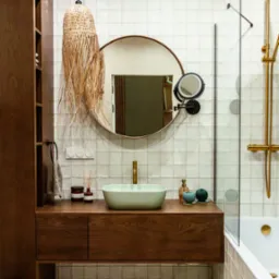 Nowoczesna łazienka z drewnianą szafką pod umywalką, okrągłym lustrem i plecioną lampą. Białe płytki z kremową fugą, złota armatura, minimalistyczny design.