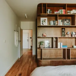 Nowoczesna sypialnia z drewnianym regałem na książki, oświetleniem punktowym i parkietem. Widok na otwarte drzwi do innego pomieszczenia. Minimalistyczny design.
