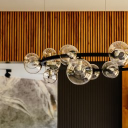 OiKa Studio Aleksandra Kośmider - Nowoczesny żyrandol z kulistymi kloszami na tle ściany z drewnianymi lamelami i dekoracyjną tapetą w odcieniach szarości, oświetlony punktowo.