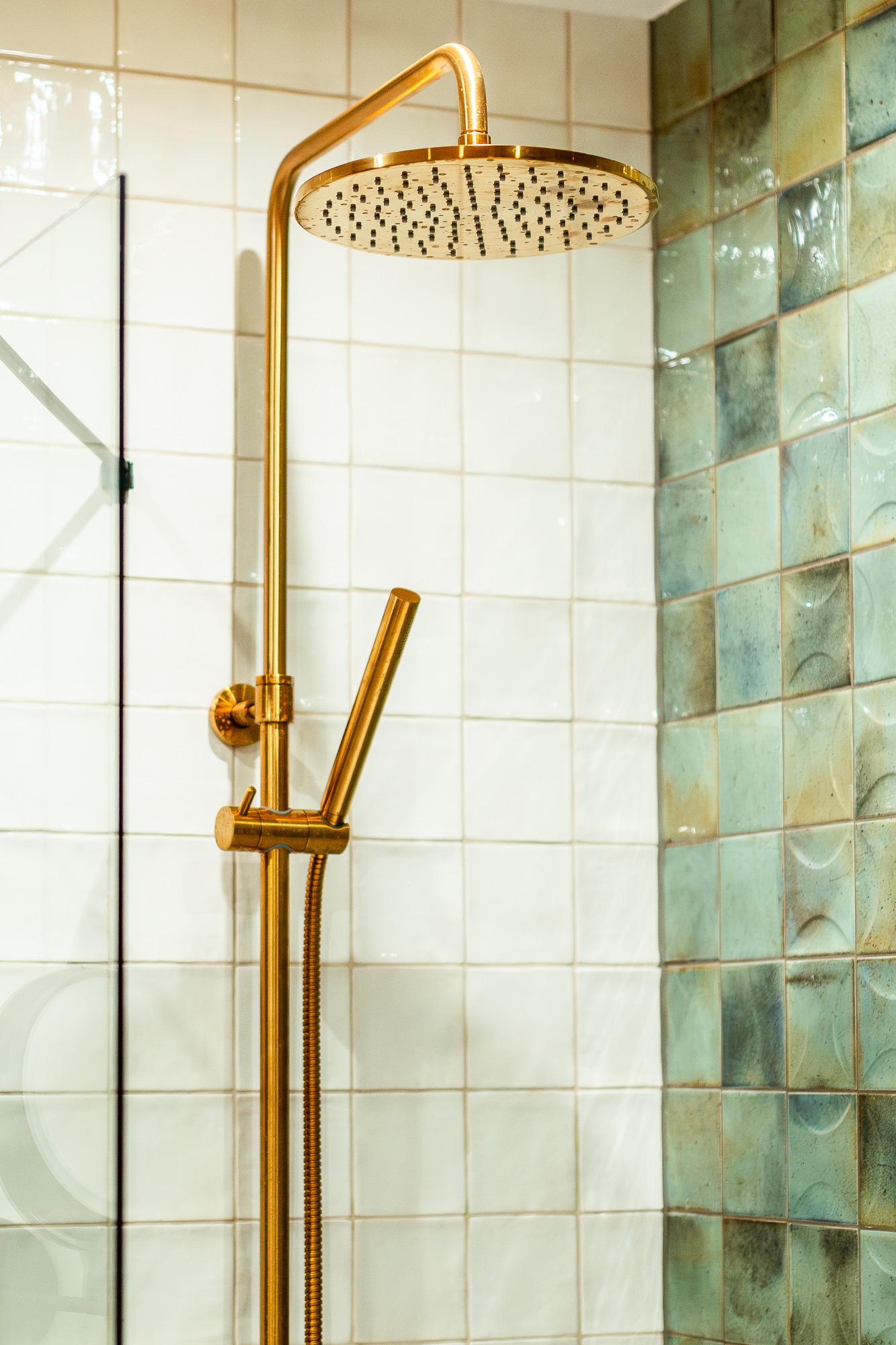 Złota bateria prysznicowa z deszczownicą na tle białych i turkusowych płytek w łazience. Widoczny fragment szklanego parawanu.