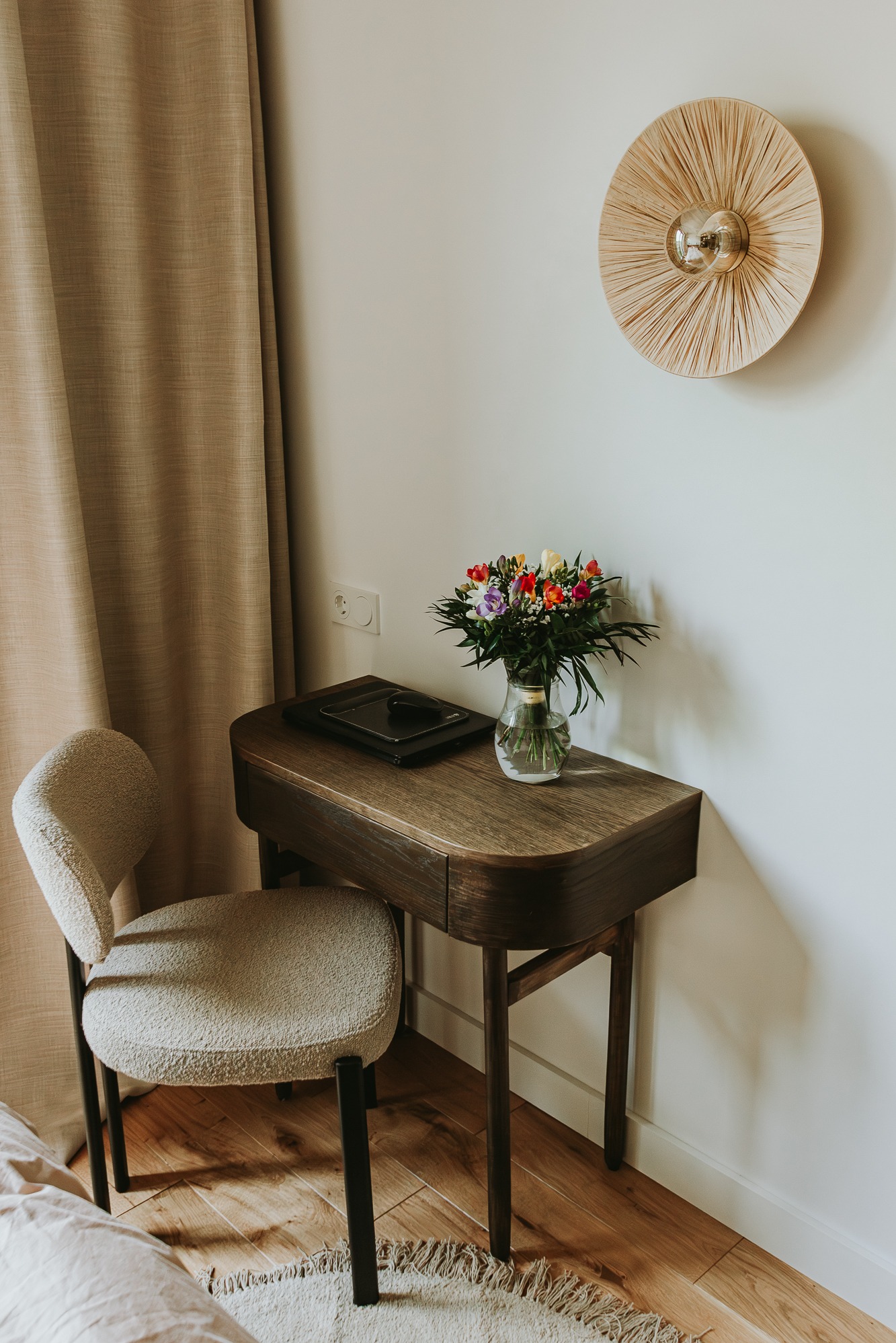 Aranżacja wnętrza: drewniane biurko z kwiatami, krzesło boucle i okrągła lampa na ścianie tworzą przytulny kącik do pracy lub relaksu. Neutralna kolorystyka.