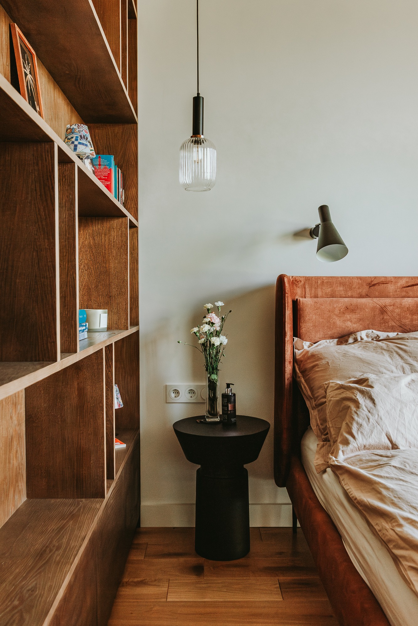 Nowoczesna sypialnia: drewniany regał, lampa wisząca, stolik nocny z kwiatami, łóżko z pomarańczowym zagłówkiem i lampką ścienną. Parkiet na podłodze.