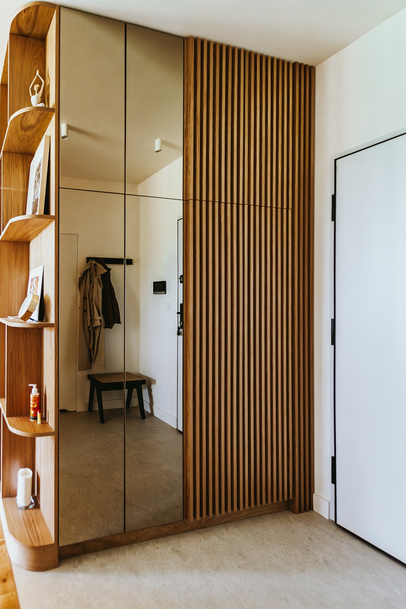 Nowoczesna szafa w przedpokoju z lustrzanymi drzwiami i drewnianymi lamelami. Na półkach dekoracje i kosmetyki. W lustrze widoczny wieszak i siedzisko.