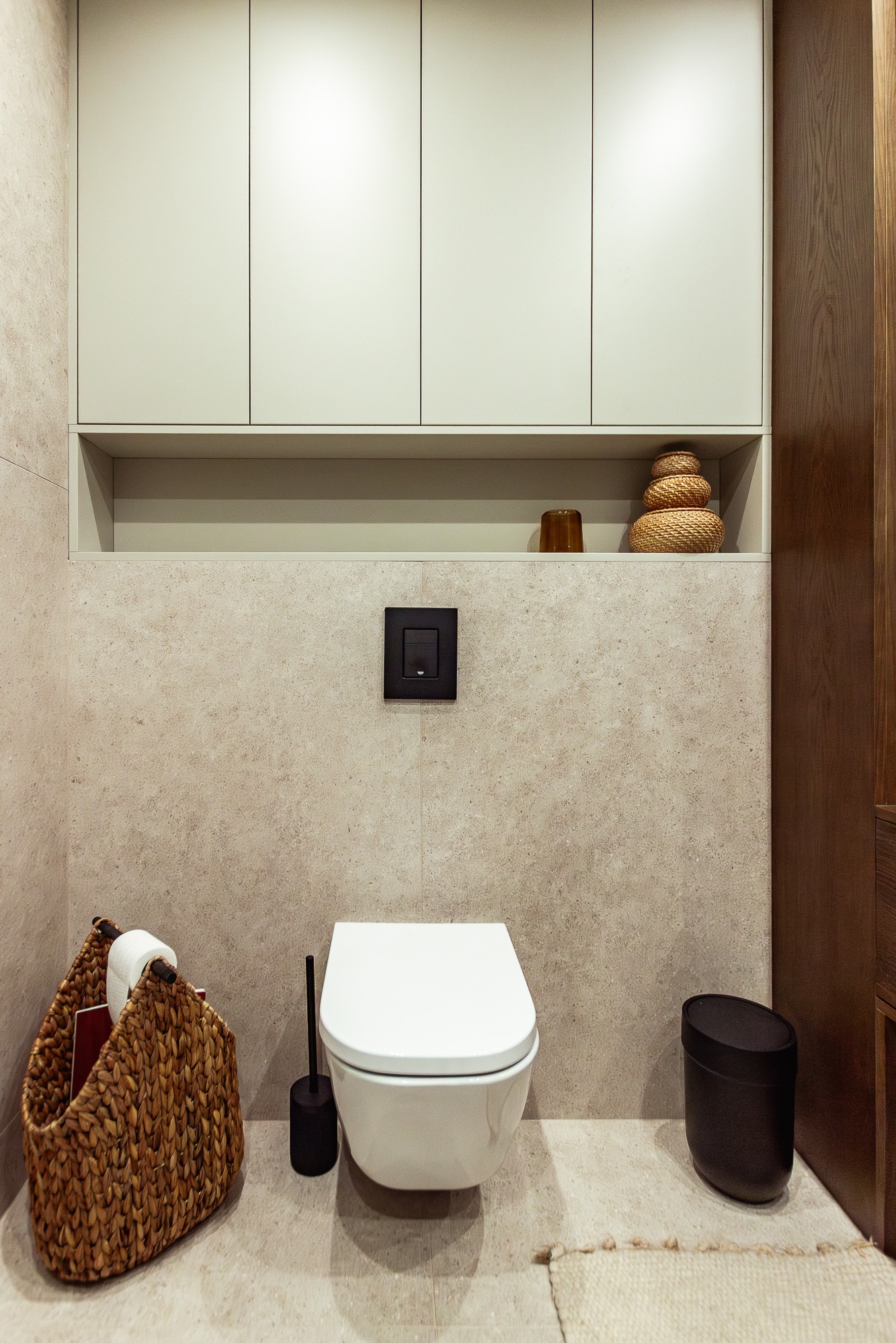 Nowoczesna toaleta podwieszana w jasnej łazience w Olkuszu. Nad nią szafki i półka z dekoracjami. Kosz z wikliny i czarne akcesoria.