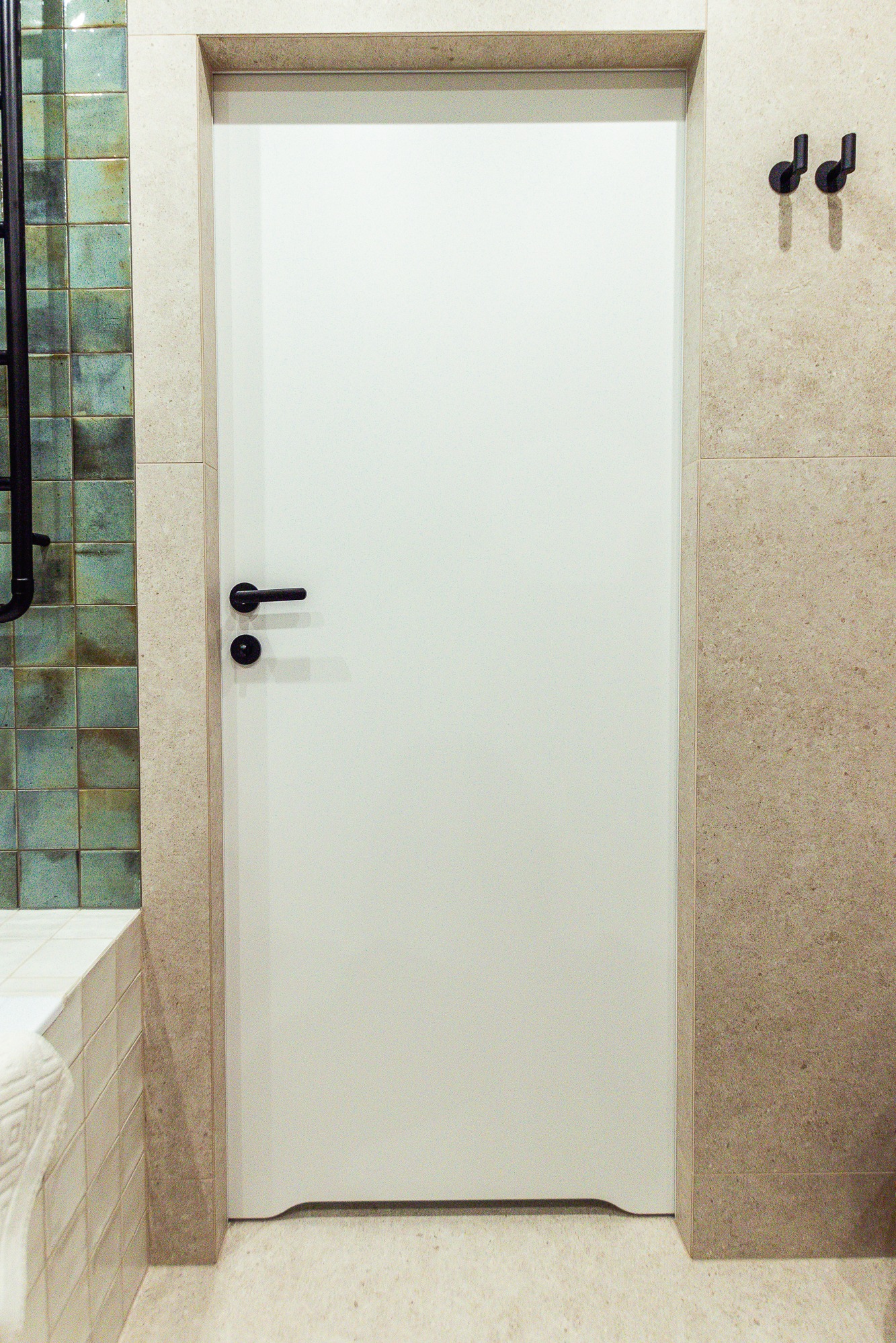 Minimalistyczna, biała łazienkowa z czarną klamką, wkomponowana w beżowe płytki. Z lewej strony widoczne turkusowe akcenty i fragment grzejnika.