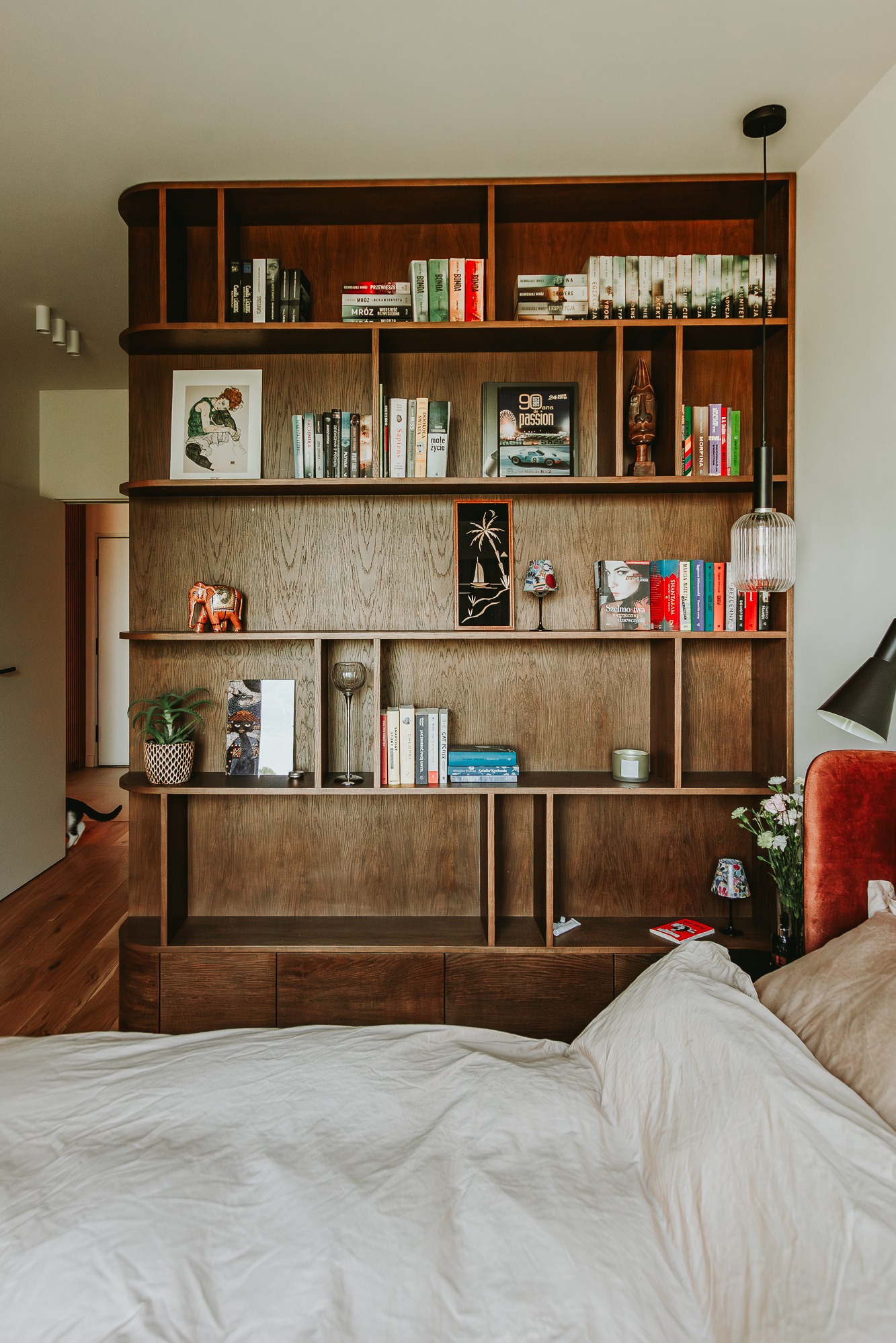 Drewniany regał z książkami i dekoracjami w sypialni, widok z perspektywy łóżka. Ciepłe oświetlenie, minimalistyczny design, elementy dekoracyjne jak figurka słonia.