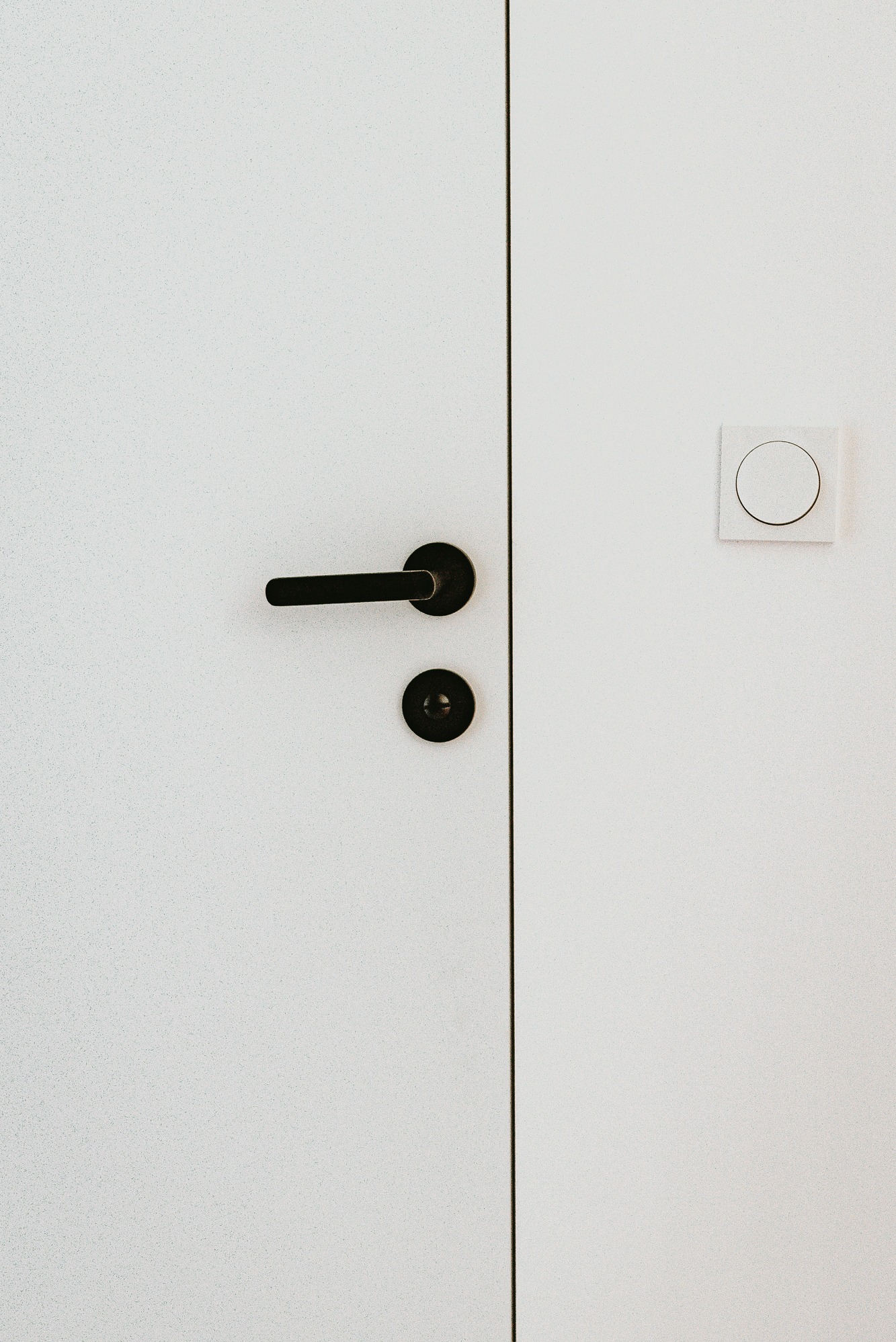 Minimalistyczne ujęcie białych drzwi z czarną klamką i okrągłym włącznikiem światła. Nowoczesny design z dbałością o detale i prostotę formy.