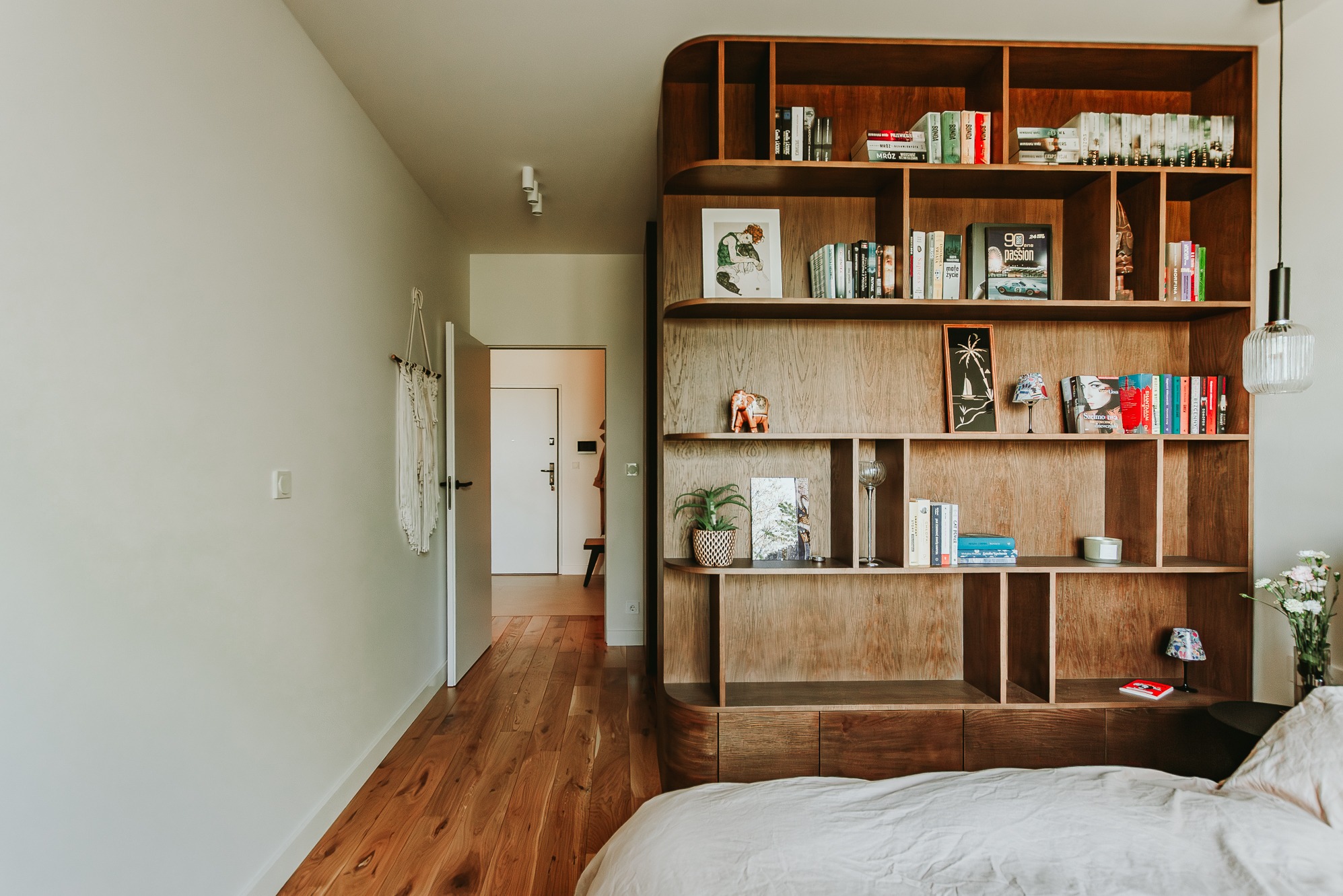 Nowoczesna sypialnia z drewnianym regałem na książki, oświetleniem punktowym i parkietem. Widok na otwarte drzwi do innego pomieszczenia. Minimalistyczny design.