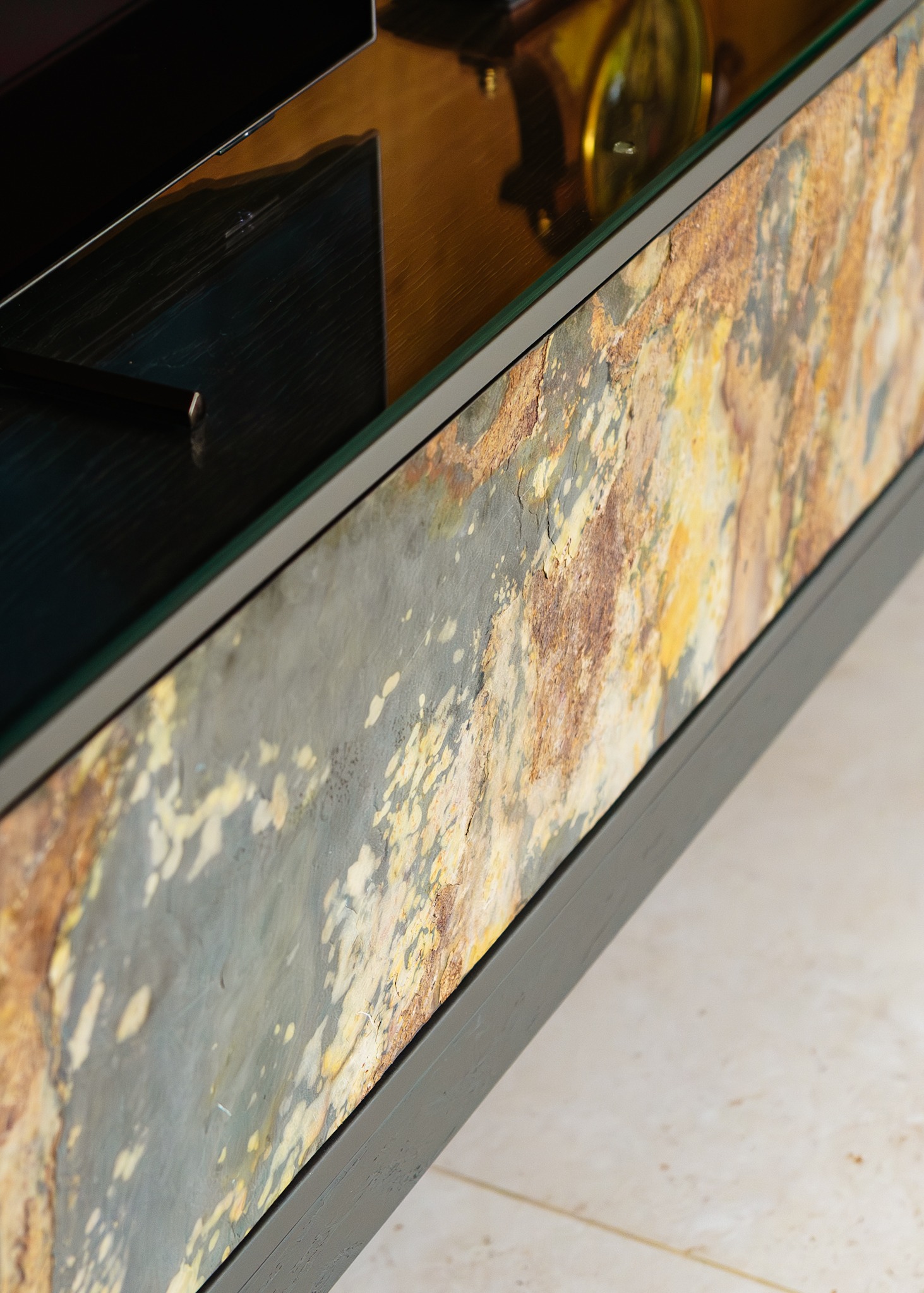 Fragment szafki RTV z blatem ze szkła i frontem imitującym kamień w odcieniach szarości i brązu. Widoczny telewizor i dekoracyjny zegar.