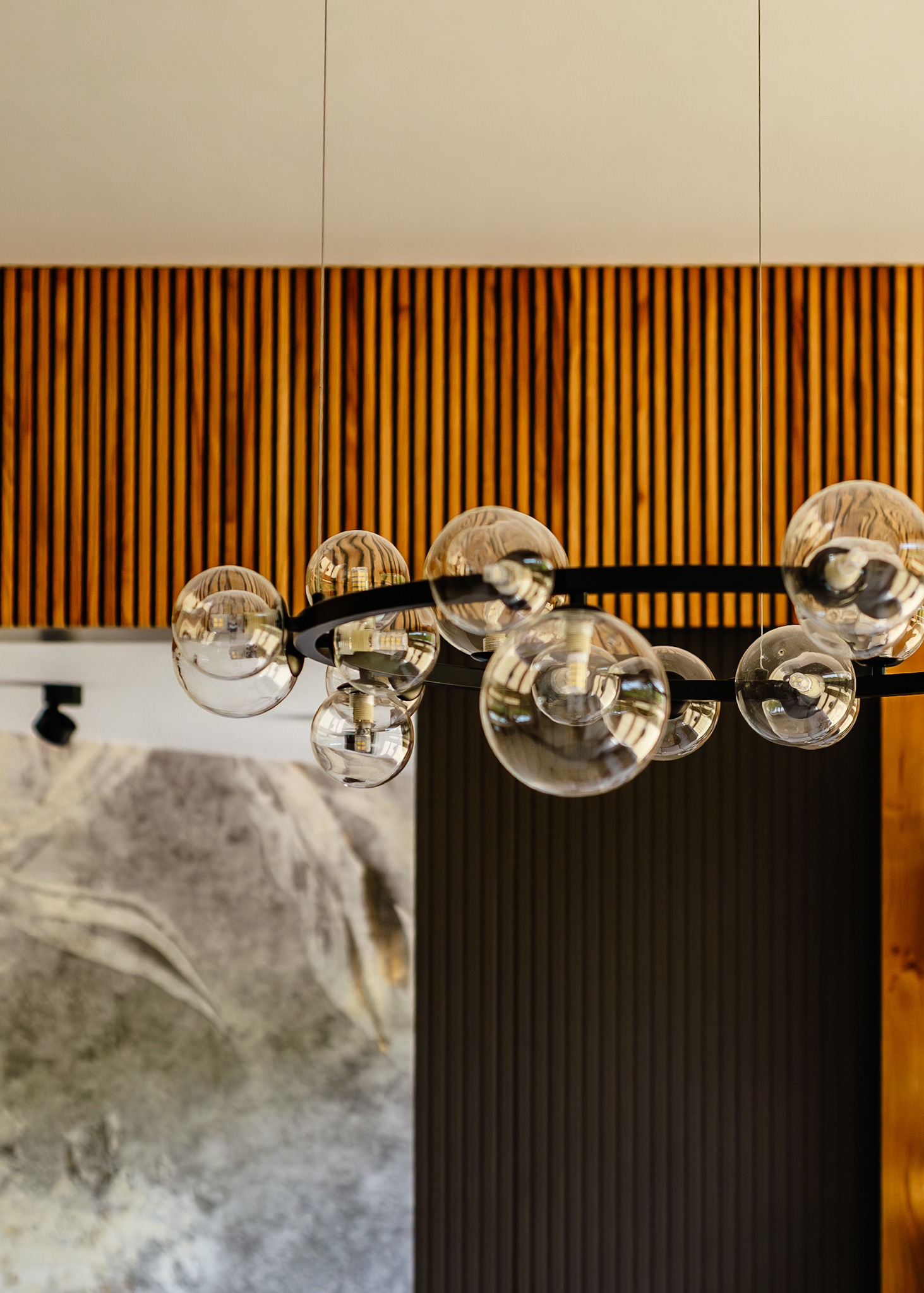 Nowoczesny żyrandol z kulistymi kloszami na tle ściany z drewnianymi lamelami i dekoracyjną tapetą w odcieniach szarości, oświetlony punktowo.