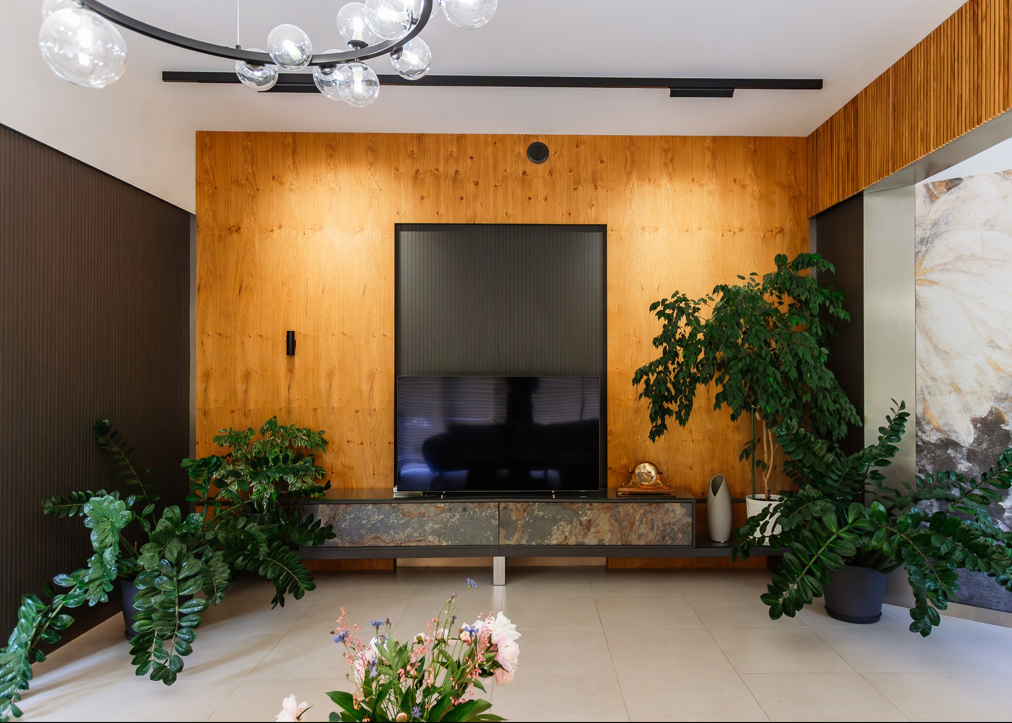 Salon z drewnianą ścianą, telewizorem na szafce z kamiennymi frontami i roślinami doniczkowymi. Nowoczesna lampa sufitowa. Jasna podłoga z płytek.