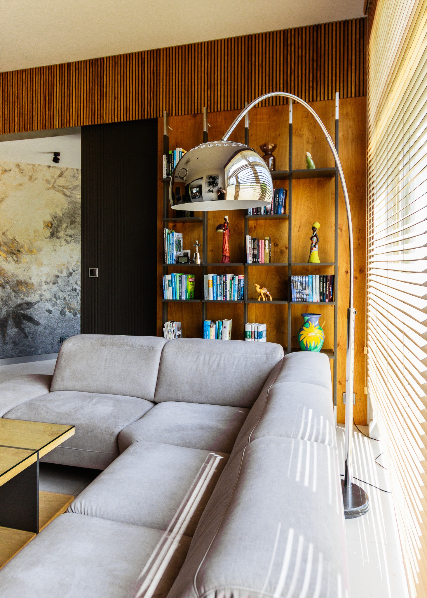 Nowoczesny salon z szarą sofą narożną, drewnianą zabudową z półkami i lampą łukową. Widoczne żaluzje w oknie i dekoracyjna ściana.