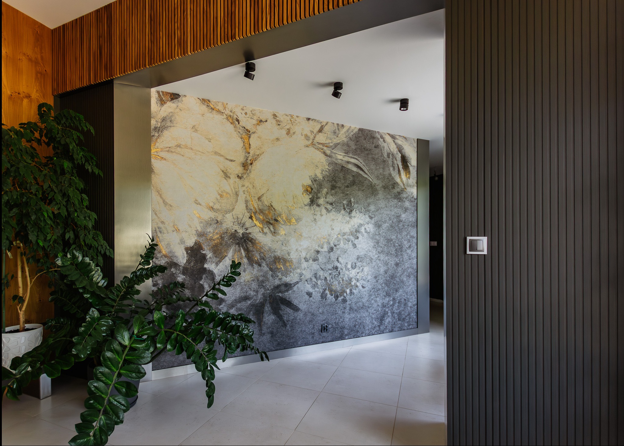 Wnętrze z dekoracyjną ścianą w odcieniach szarości i złota, oświetleniem punktowym. W rogu widoczna zielona roślina doniczkowa, a obok ściana z paneli pionowych.