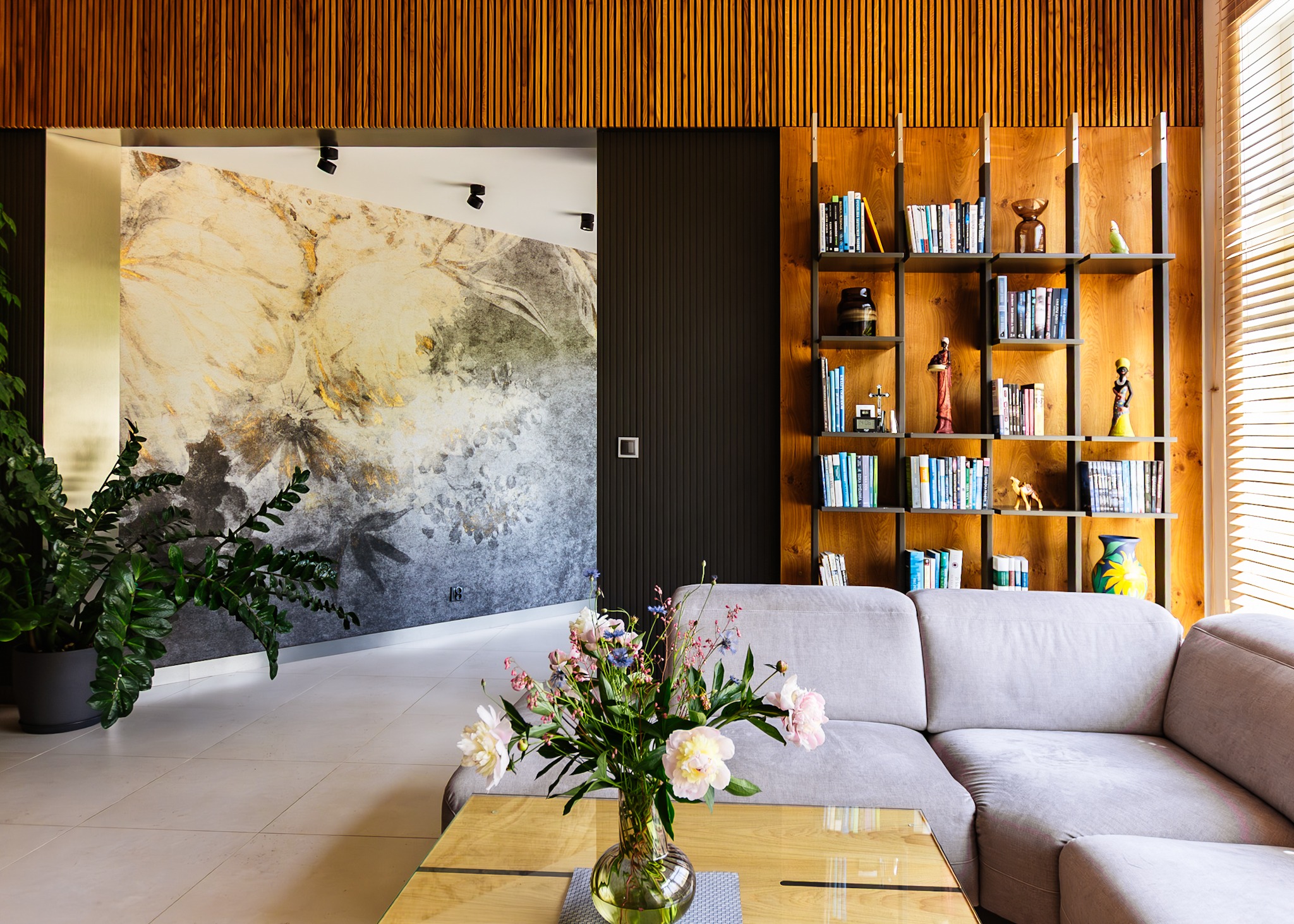 Nowoczesny salon z kwiatami w wazonie na stoliku, szarą sofą i regałem z książkami na tle drewnianej ściany. Na ścianie abstrakcyjny wzór z motywem kwiatowym.