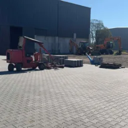 Maszyna do układania kostki brukowej na tle hali, ułożona kostka, palety z kostką, zagęszczarka i koparki, błękitne niebo.
