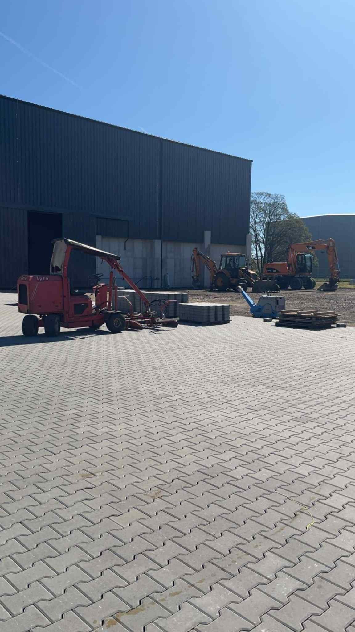Maszyna do układania kostki brukowej na tle hali, ułożona kostka, palety z kostką, zagęszczarka i koparki, błękitne niebo.