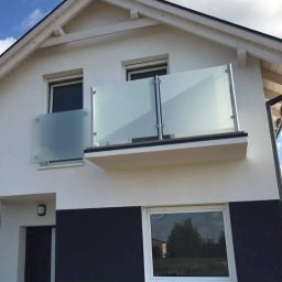 NAJDEK PRO SPÓŁKA Z OGRANICZONĄ ODPOWIEDZIALNOŚCIĄ - Nowoczesna balustrada szklana z aluminiowymi słupkami, zamontowana na balkonie domu z biało-czarną elewacją. Widok z dołu, błękitne niebo w tle.