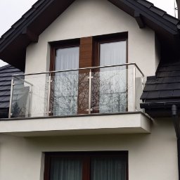 NAJDEK PRO SPÓŁKA Z OGRANICZONĄ ODPOWIEDZIALNOŚCIĄ - Balkon z nowoczesną, szklaną balustradą ze stali nierdzewnej na tle jasnej elewacji domu z ciemnym dachem. Widok z dołu, odbicie drzew w szkle.