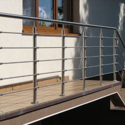 NAJDEK PRO SPÓŁKA Z OGRANICZONĄ ODPOWIEDZIALNOŚCIĄ - Błyszcząca, metalowa balustrada z poziomymi prętami na balkonie z brązowymi płytkami, przy jasnej ścianie budynku z oknem i zielenią w tle.