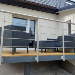 Balkon z drewnianą podłogą i stalową balustradą z okrągłych profili. Na balkonie stoją rattanowe meble wypoczynkowe w kolorze grafitowym.
