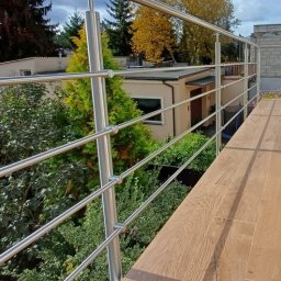 NAJDEK PRO SPÓŁKA Z OGRANICZONĄ ODPOWIEDZIALNOŚCIĄ - Nowoczesna, trzypoziomowa konstrukcja ze stali nierdzewnej w Szubinie. Widok z góry na drewnopodobną podłogę balkonu i zieleń w tle. Słoneczny dzień.