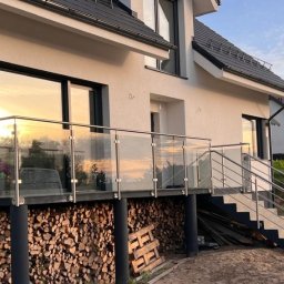 NAJDEK PRO SPÓŁKA Z OGRANICZONĄ ODPOWIEDZIALNOŚCIĄ - Nowoczesna balustrada szklana z elementami ze stali nierdzewnej na balkonie domu jednorodzinnego z widokiem na zachód słońca odbijającym się w szybach.