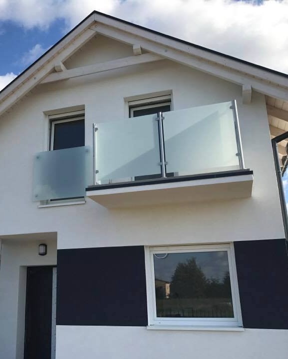 Nowoczesna balustrada szklana z aluminiowymi słupkami, zamontowana na balkonie domu z biało-czarną elewacją. Widok z dołu, błękitne niebo w tle.