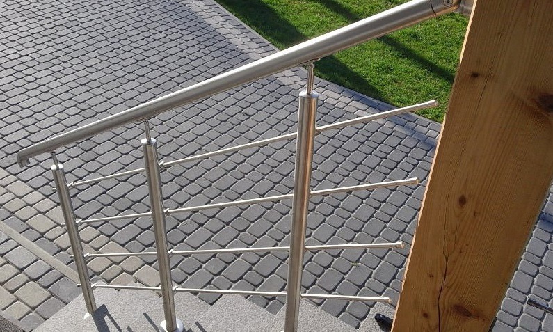 Metalowa balustrada ze szczeblami, widok z góry na schody wyłożone kostką. Elementy drewniane w tle. Jasne światło dzienne.