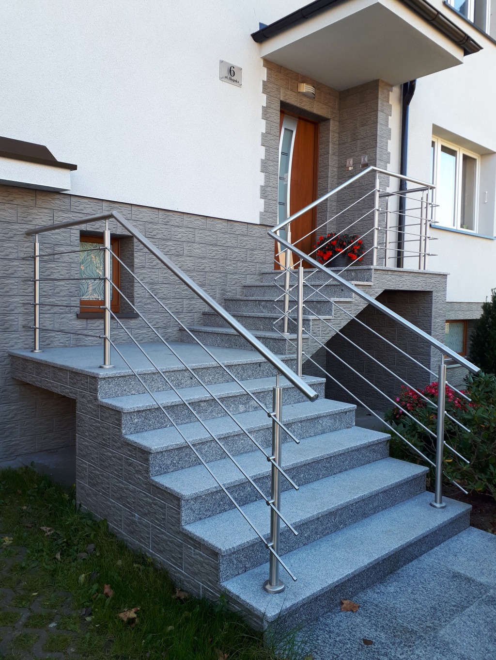 Zewnętrzne schody z szarego granitu prowadzą do drzwi domu. Uwagę przyciągają nowoczesne, poziome balustrady ze stali nierdzewnej z delikatnymi kwiatami w tle.