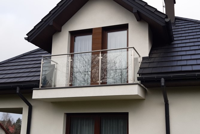 Balkon z nowoczesną, szklaną balustradą ze stali nierdzewnej na tle jasnej elewacji domu z ciemnym dachem. Widok z dołu, odbicie drzew w szkle.