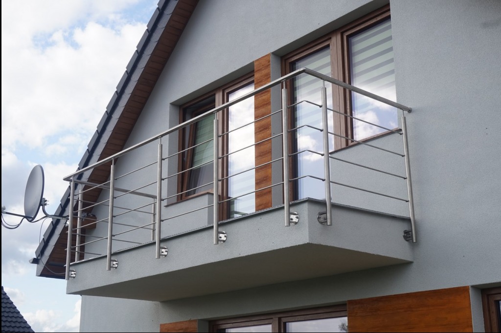 Nowoczesny balkon z szarą elewacją i balustradą ze stali nierdzewnej, widok na okna z drewnianymi ramami i częściowo widoczną anteną satelitarną.