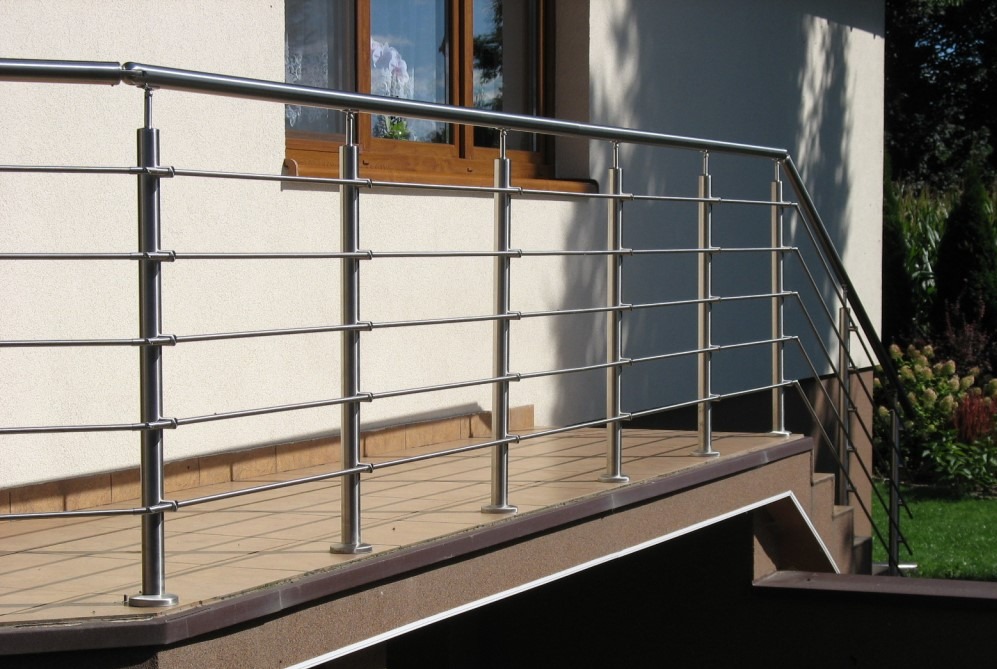 Błyszcząca, metalowa balustrada z poziomymi prętami na balkonie z brązowymi płytkami, przy jasnej ścianie budynku z oknem i zielenią w tle.