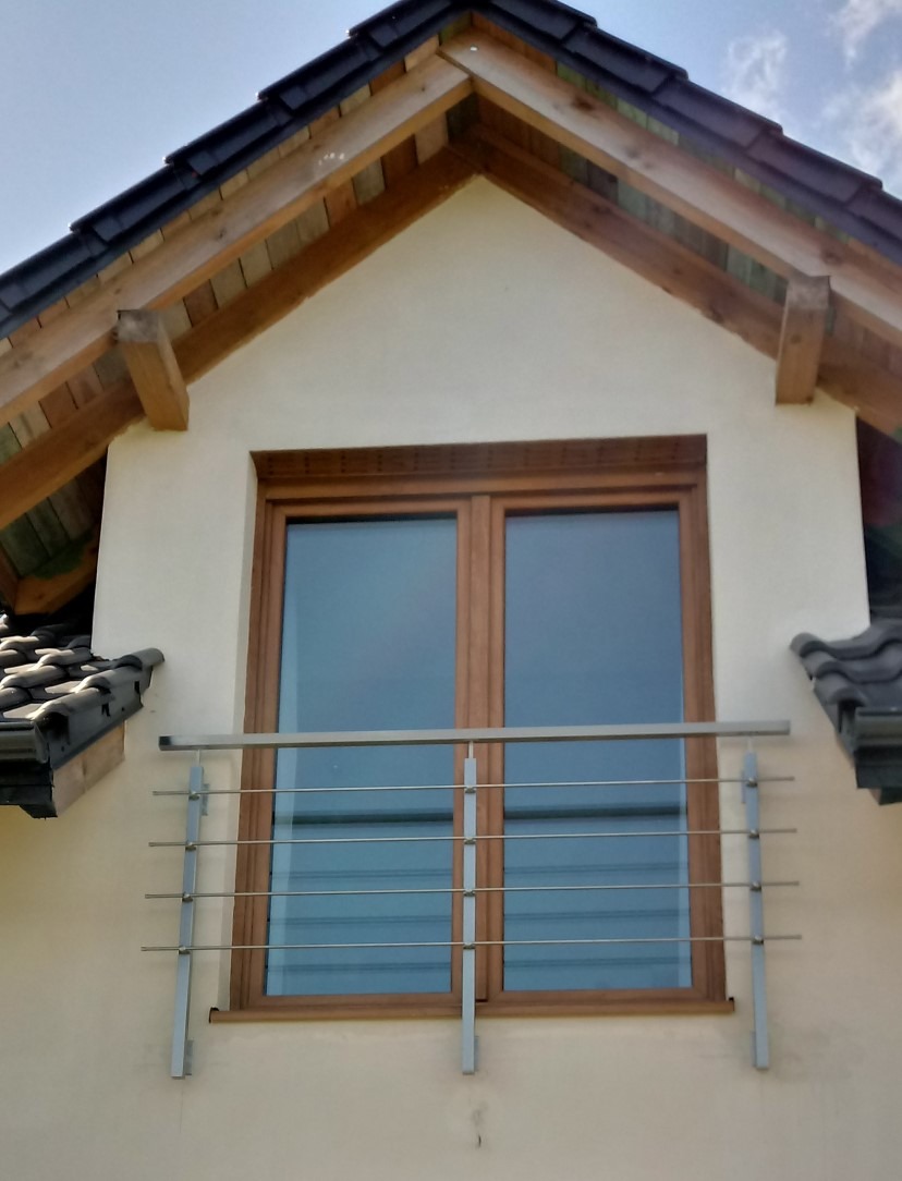 Nierdzewna balustrada okienna z poziomymi prętami, zamontowana na elewacji domu z drewnianą stolarką okienną i ciemną dachówką. Detal architektoniczny.