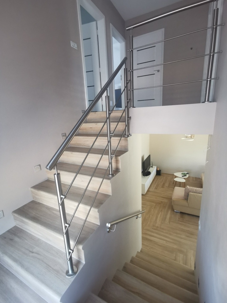 Nowoczesne schody z drewnianymi stopniami i minimalistyczną balustradą ze stali nierdzewnej. Widok na salon z parkietem w jodełkę i beżową sofą.