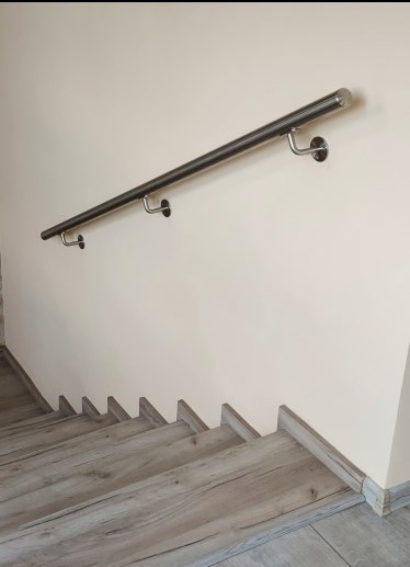 Nierdzewna poręcz zamontowana na ścianie przy schodach z drewnopodobnymi panelami. Minimalistyczny design, jasne wnętrze.