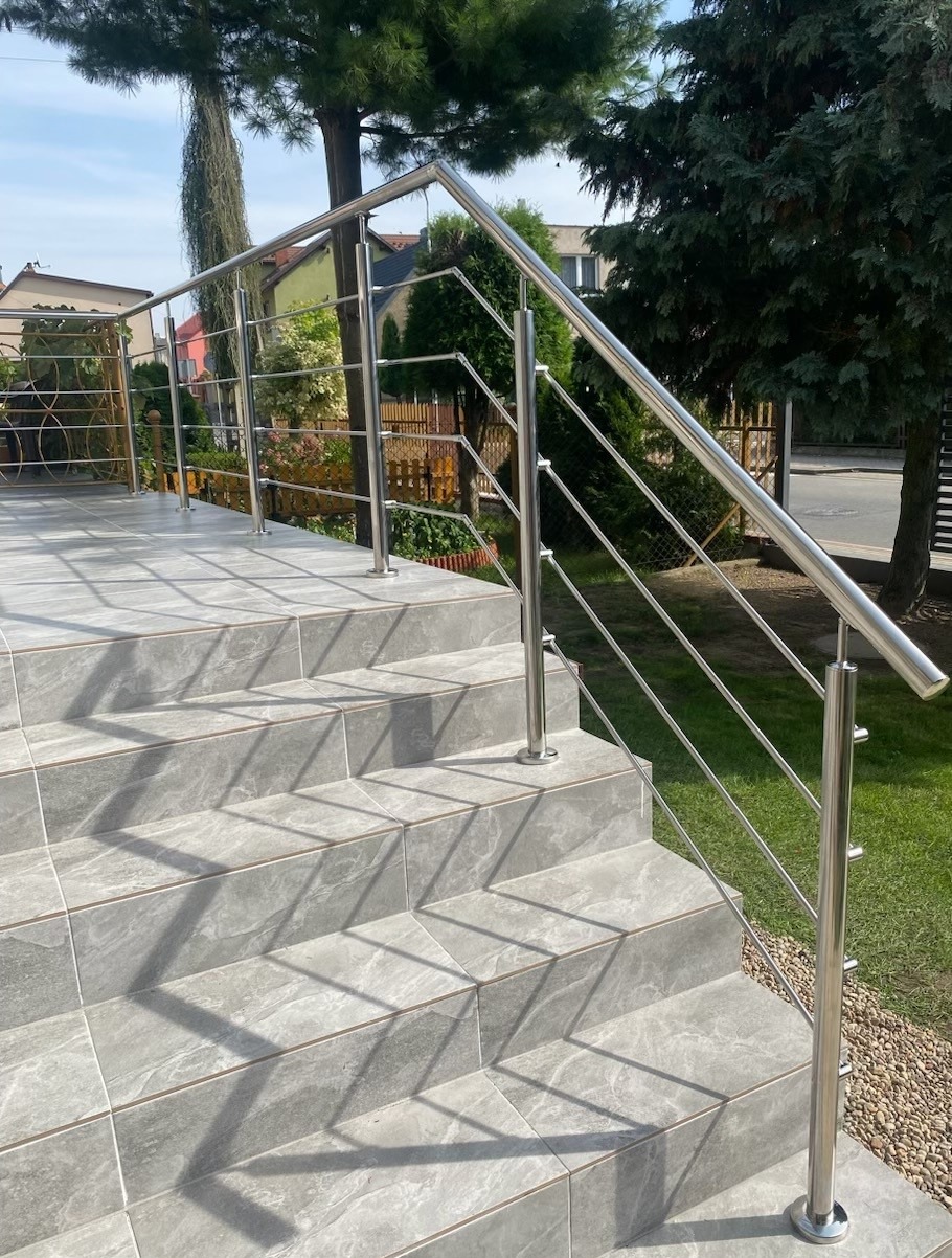Polerowana, stalowa balustrada schodowa z poziomymi prętami, zamontowana na zewnątrz budynku z szarymi płytkami. Widok na zielony trawnik i drzewa.