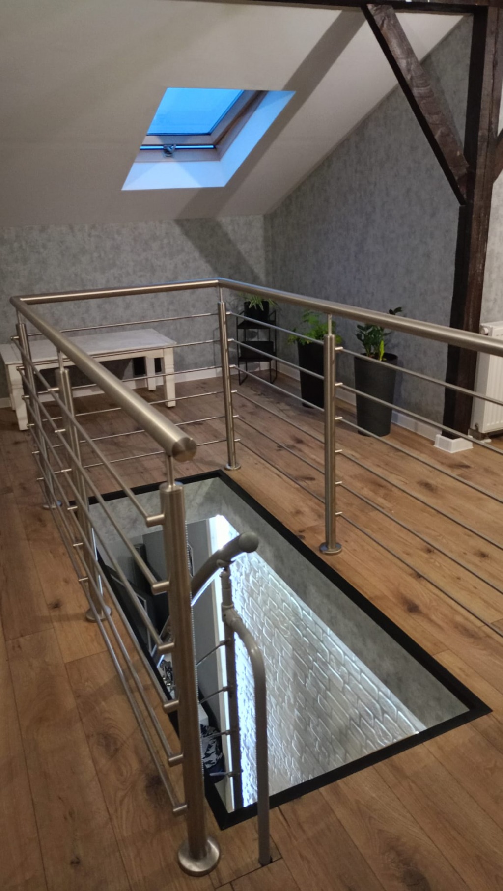 Nietypowa balustrada ze stali nierdzewnej z poziomymi prętami, zabezpieczająca otwór w podłodze. Widok z góry, drewniana podłoga i okno dachowe.
