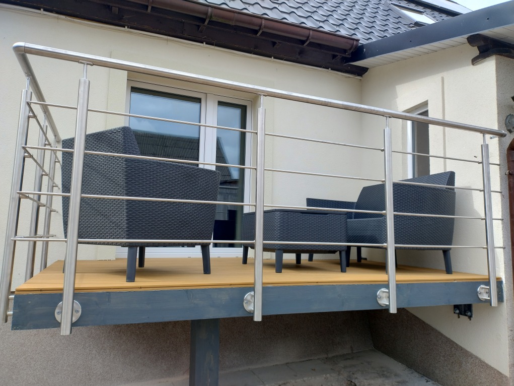 Balkon z drewnianą podłogą i stalową balustradą z okrągłych profili. Na balkonie stoją rattanowe meble wypoczynkowe w kolorze grafitowym.