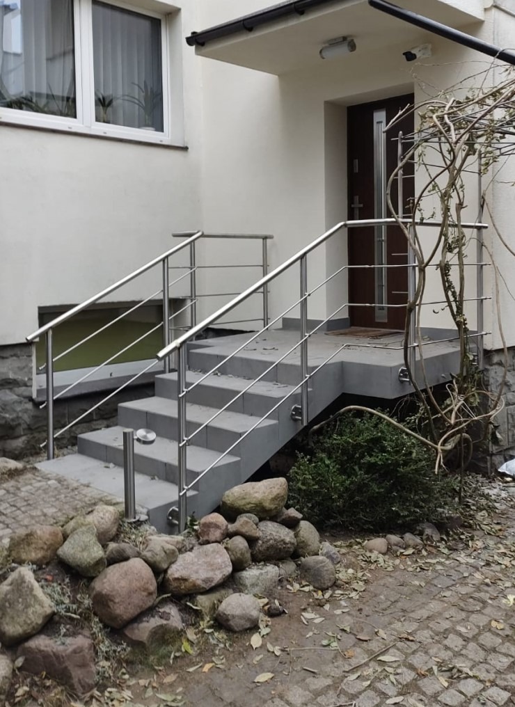 Zewnętrzne schody z nowoczesnymi, srebrnymi poręczami prowadzące do drzwi wejściowych. Widoczny fragment fasady budynku i kamienny ogródek.