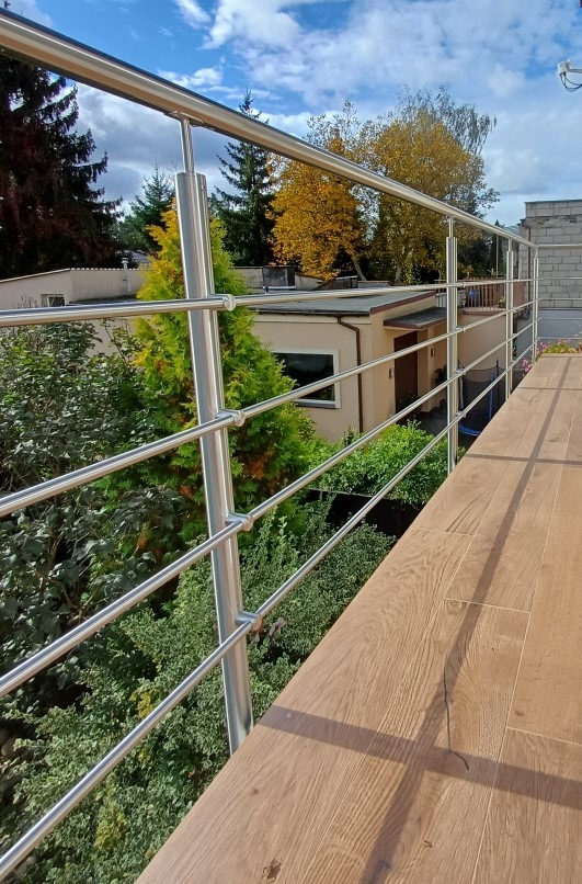 Nowoczesna, trzypoziomowa konstrukcja ze stali nierdzewnej w Szubinie. Widok z góry na drewnopodobną podłogę balkonu i zieleń w tle. Słoneczny dzień.