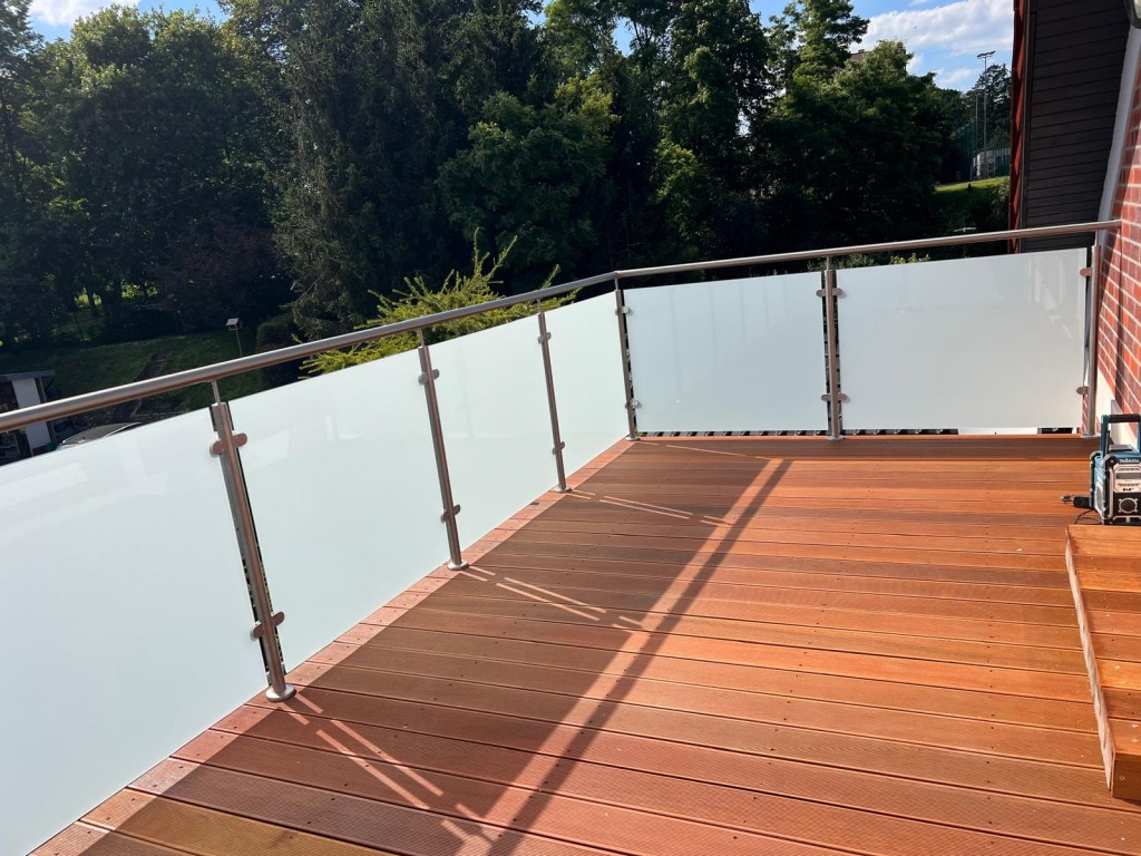 Balkon z drewnianą podłogą i balustradą ze stali nierdzewnej z mlecznym szkłem, narzędzie akumulatorowe w rogu, widok na zieleń.