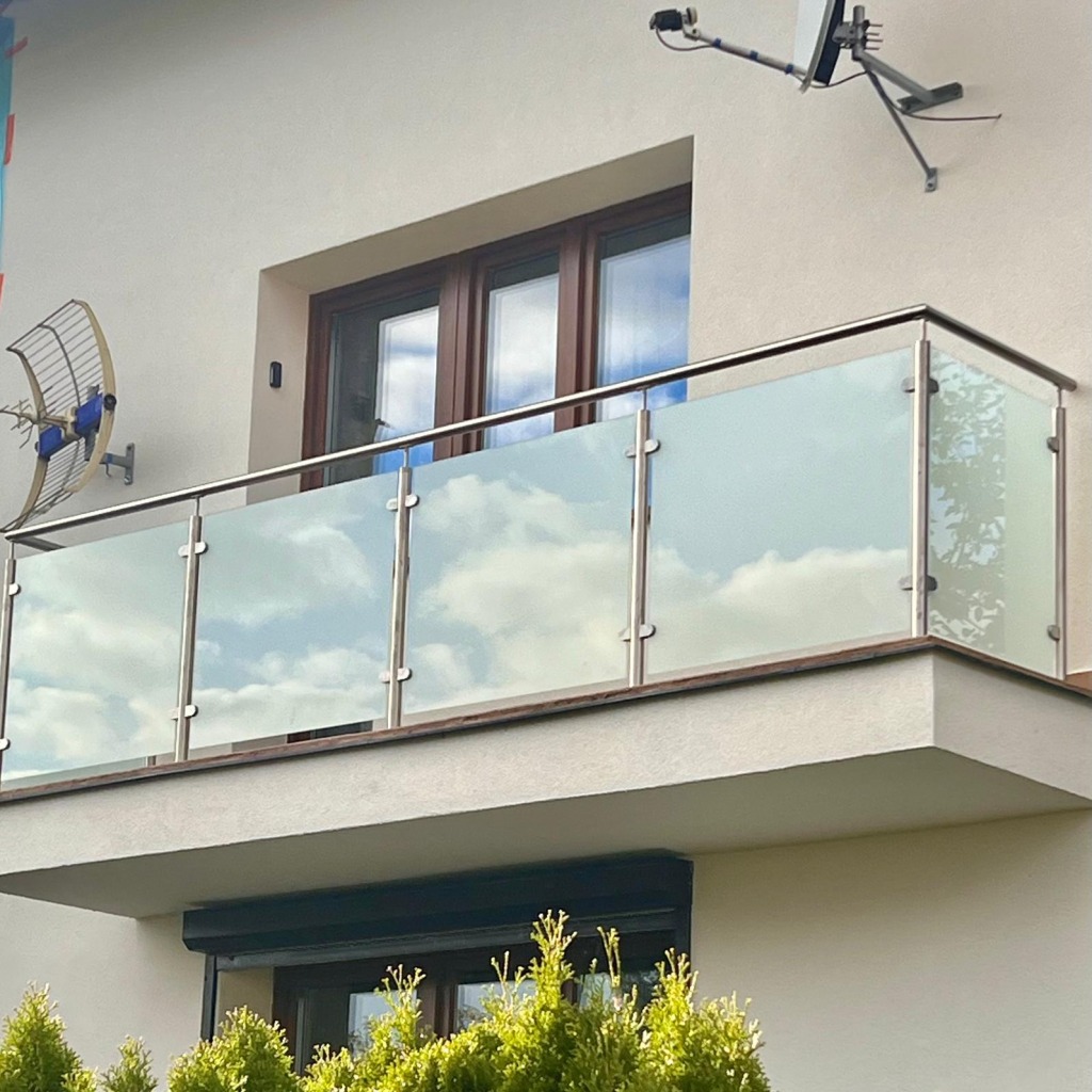 Nowoczesna balustrada ze stali nierdzewnej i mlecznego szkła na balkonie. W szkle odbijają się chmury. Na elewacji widoczna antena.