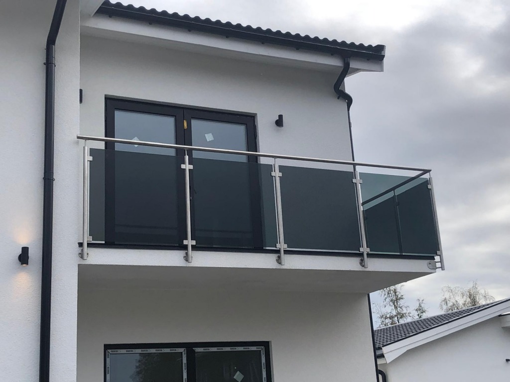 Nowoczesna balustrada balkonowa ze szkła grafitowego i stali nierdzewnej, mocowana do białej elewacji budynku z oknami. Widok z dołu.