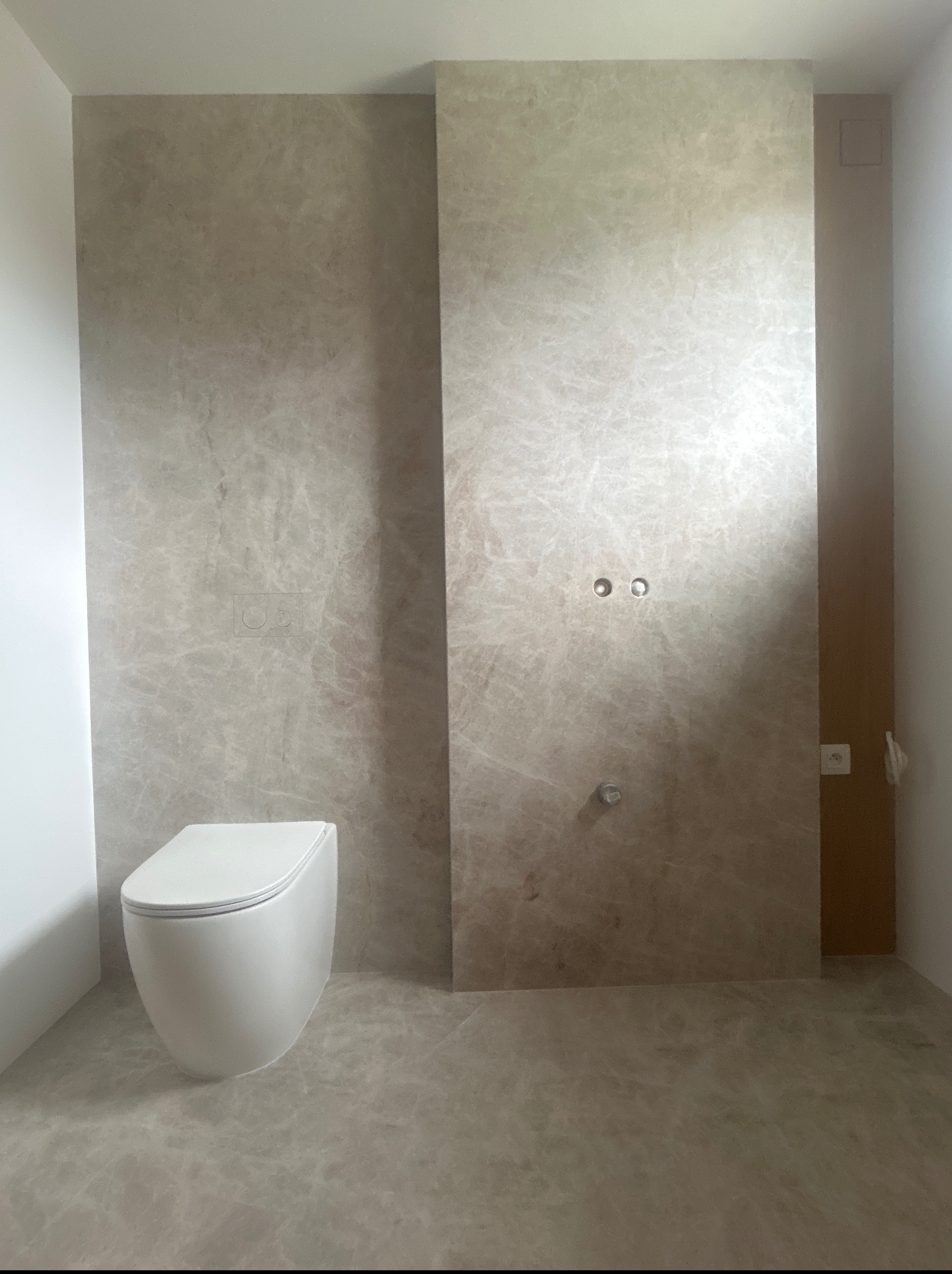 Beżowa łazienka z marmurowymi płytkami i podwieszaną toaletą. Widoczne przyłącza wodne i elektryczne, gotowe do montażu armatury. Minimalistyczny styl.