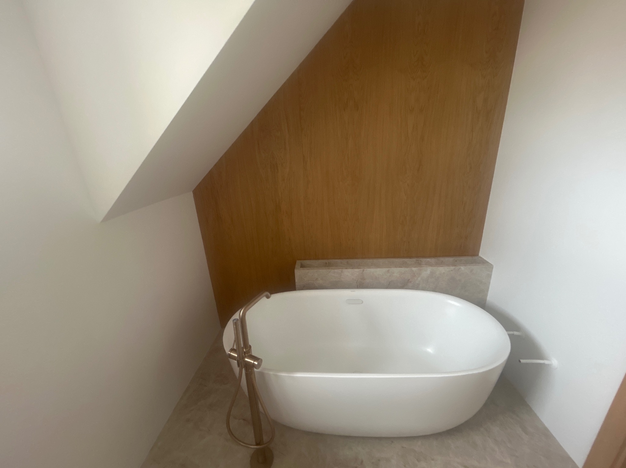 Nowoczesna, wolnostojąca wanna z złotą armaturą w jasnej łazience na poddaszu, wykończonej drewnem i marmurem. Minimalistyczny design, skupienie na detalu.