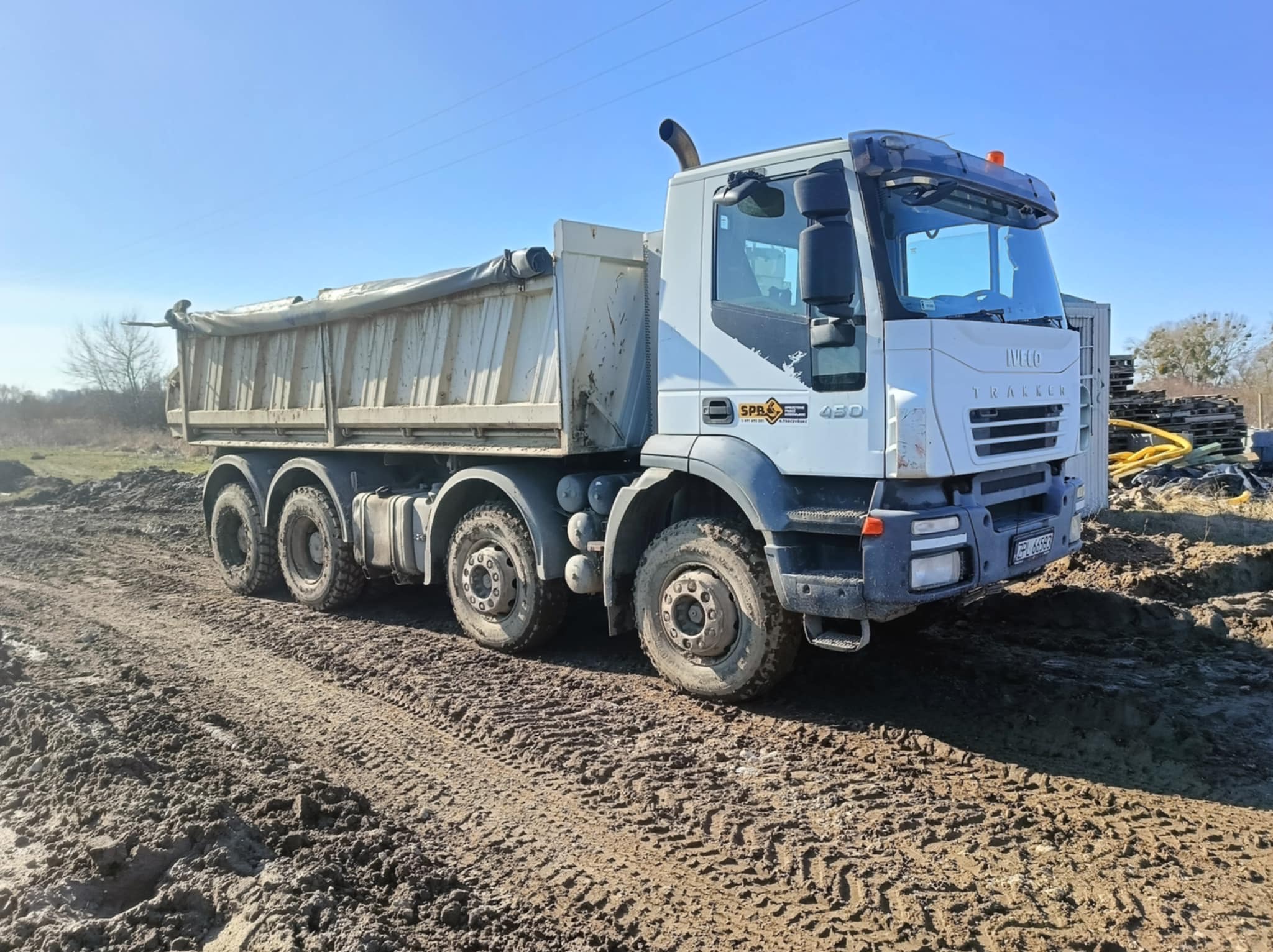 Ciężarówka Iveco Trakker 450 na błotnistym terenie, z widocznymi śladami opon i częściowo przykrytą plandeką skrzynią ładunkową, błękitne niebo w tle.
