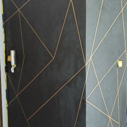 BUDMASTER - Ściana w odcieniach szarości z geometrycznym wzorem z cienkich, złotych linii. Widoczne zabezpieczenia podłogi i gniazdka elektryczne.