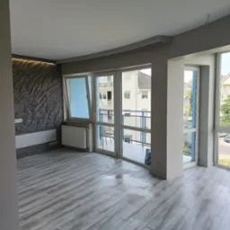 Nowoczesne wnętrze z szarą, teksturowaną ścianą, panelami podłogowymi i dużymi oknami balkonowymi z widokiem na zabudowę miejską. Minimalistyczny design i jasne, naturalne światło.