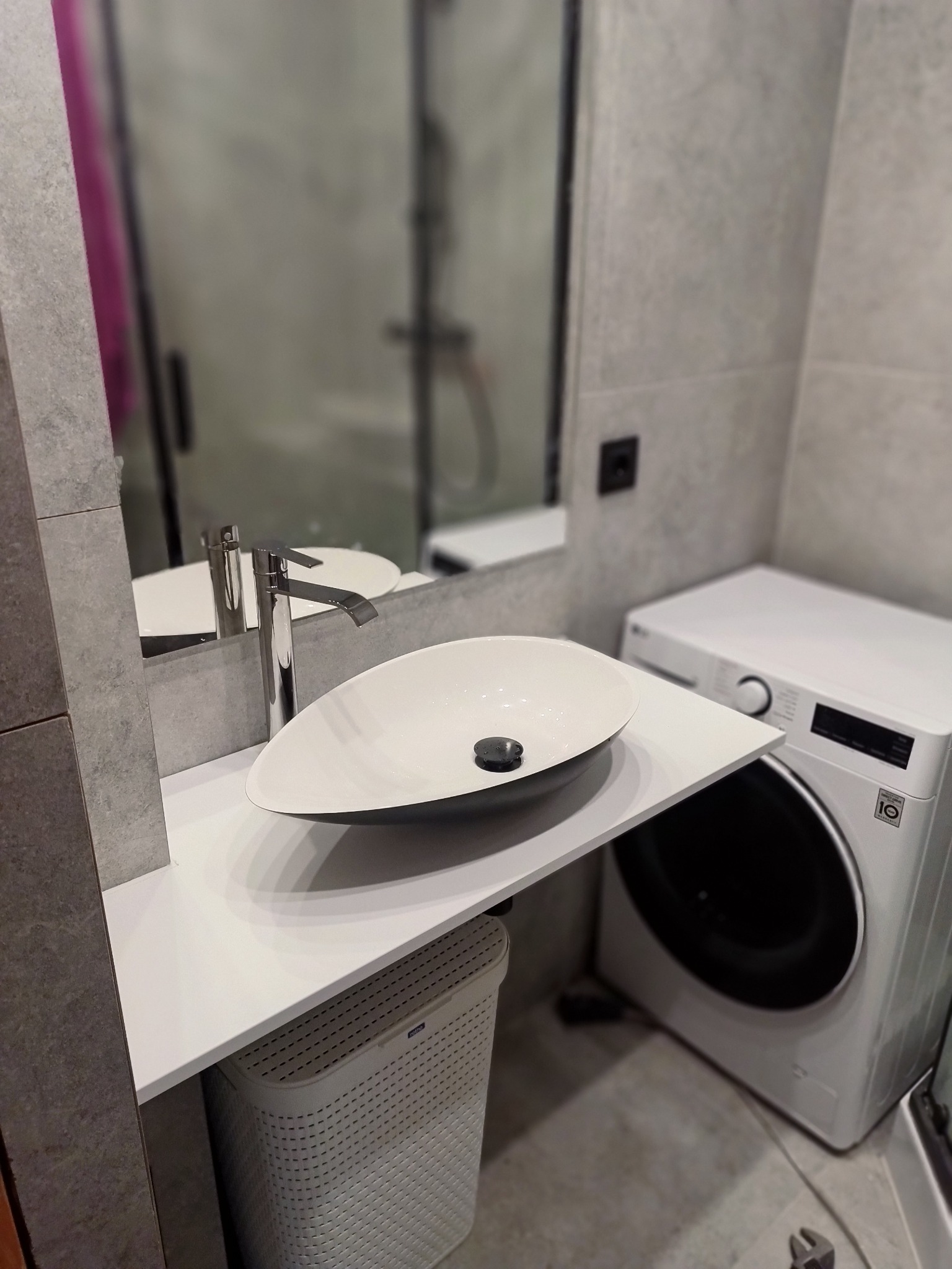 Nowoczesna łazienka z białą umywalką nablatową, chromowaną baterią i pralką LG. Minimalistyczny design, szare płytki, lustro i kosz na pranie.