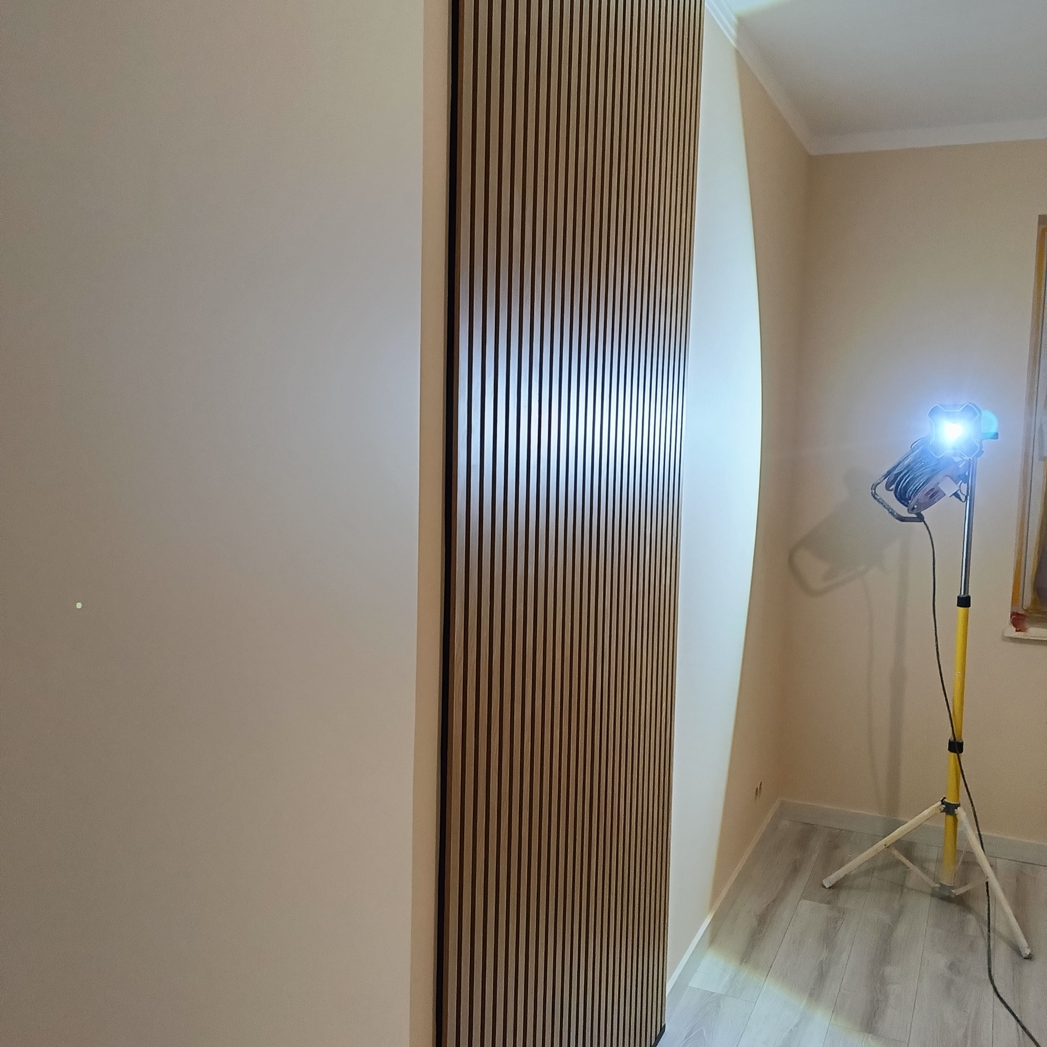 Drewniane lamele na ścianie, oświetlone lampą budowlaną. Widoczne wykończenie wnętrza z dbałością o detale i nowoczesny design.